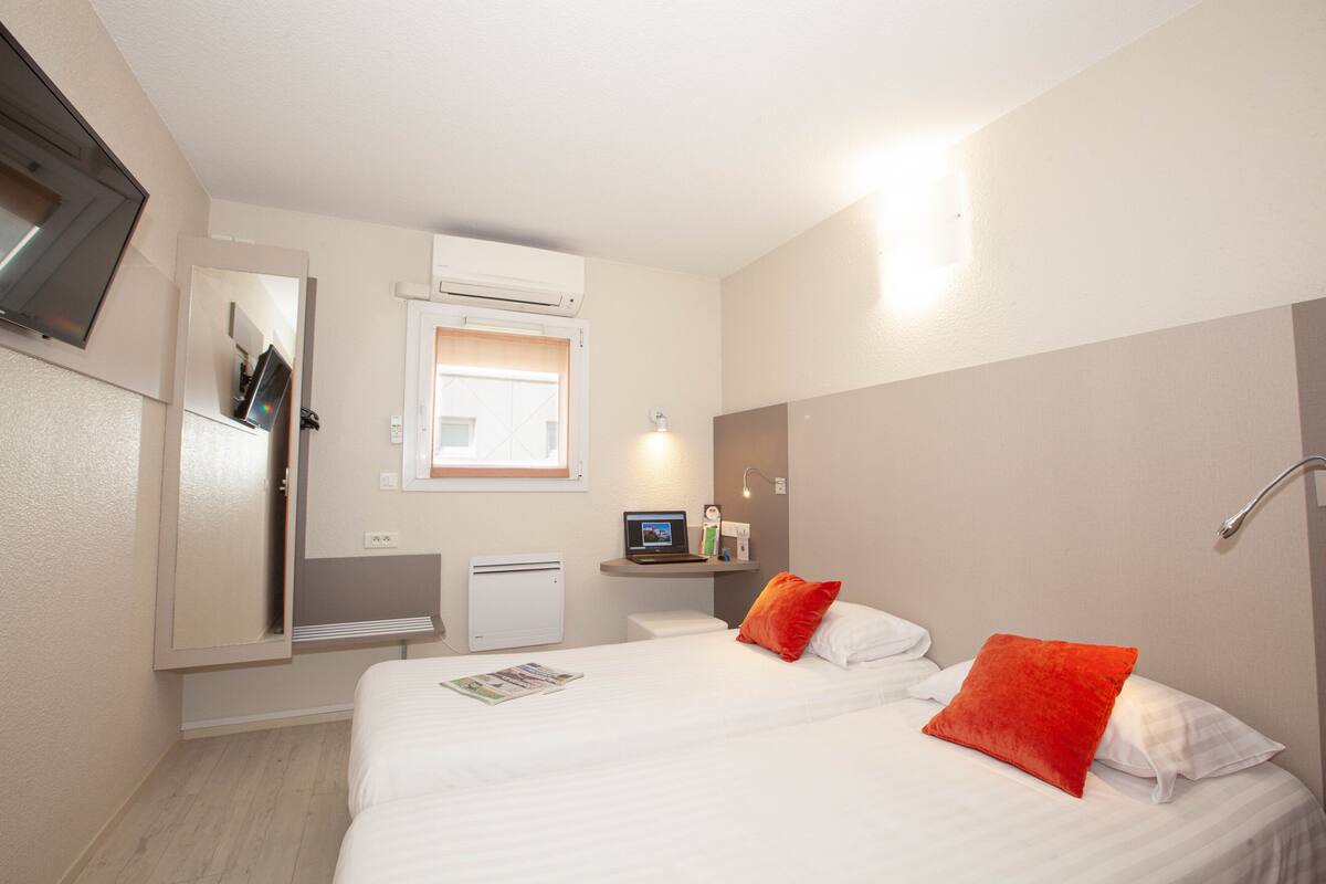 Une chambre d’hôtel avec un lit, un bureau avec un ordinateur portable, une petite étagère avec des articles de toilette et une fenêtre.
