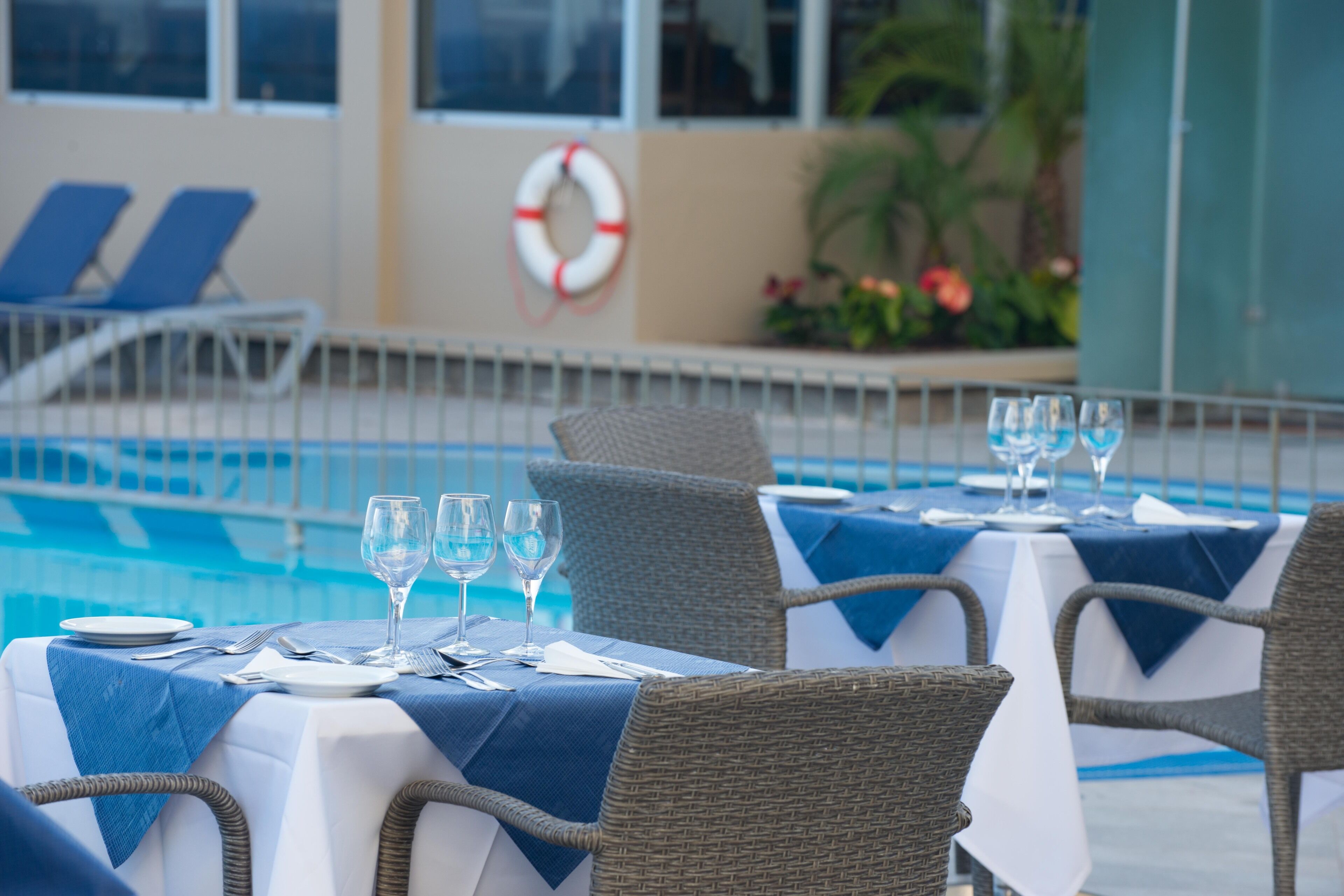A poolside dining area with tables set for two, wicker chairs, and a lifebuoy on the wall.