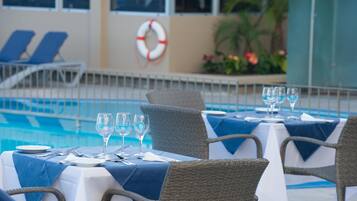 A poolside dining area with tables set for two, wicker chairs, and a lifebuoy on the wall.