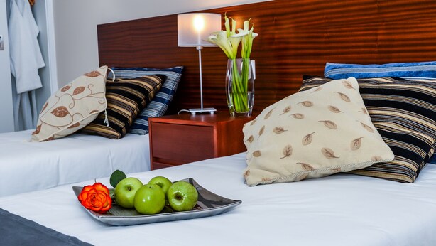 A hotel room with two beds, a bedside table with a lamp, a vase with flowers, and a tray with apples and a rose. - Hotel Duas Torres (Funchal)