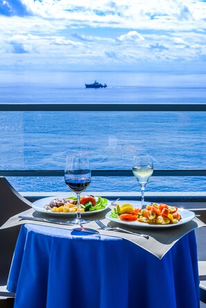 A table with two plates of food and two glasses of wine, set against a view of the ocean and a ship in the distance.