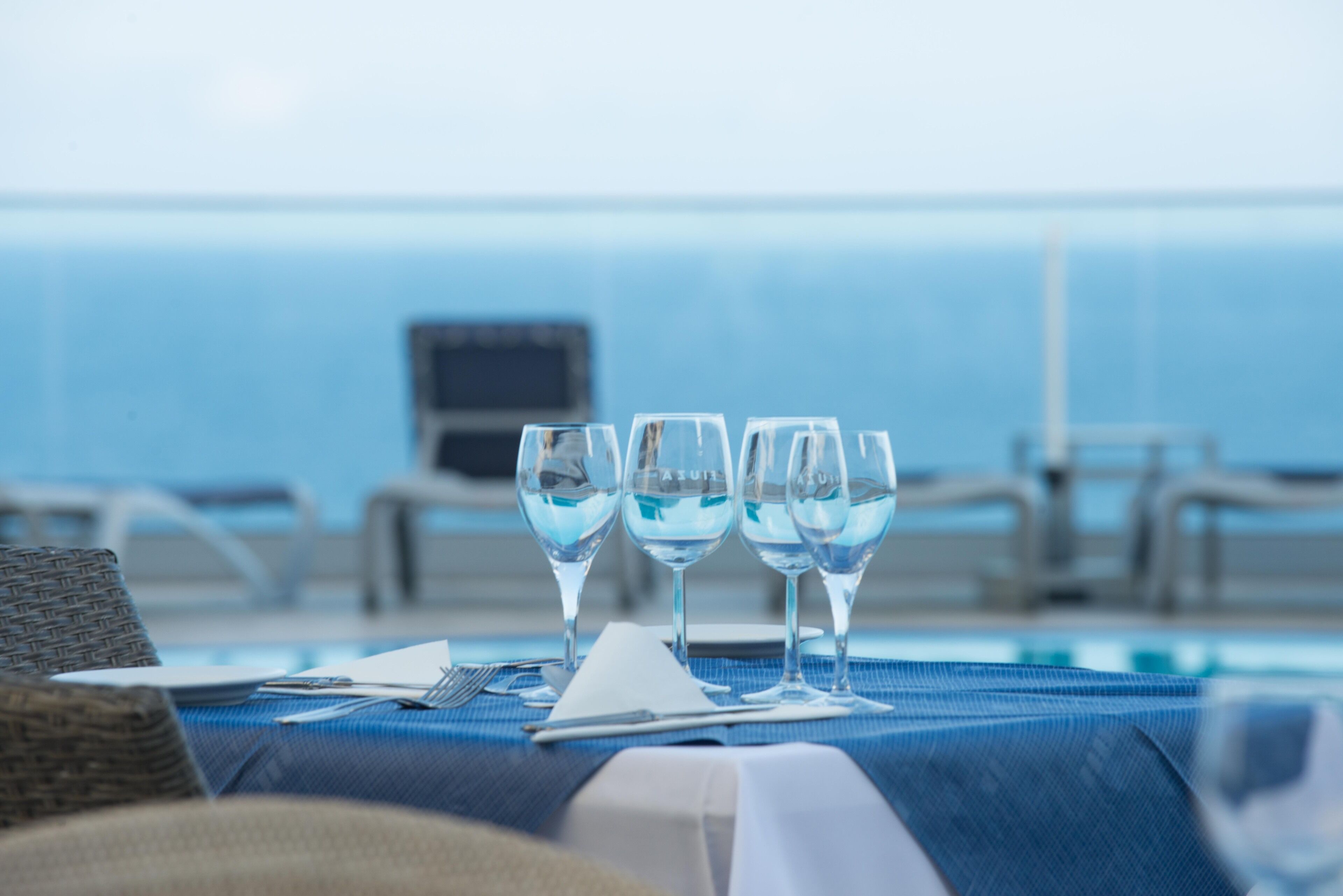 A table with four wine glasses, a laptop, and a notepad on a blue tablecloth by a pool with a view of the ocean.