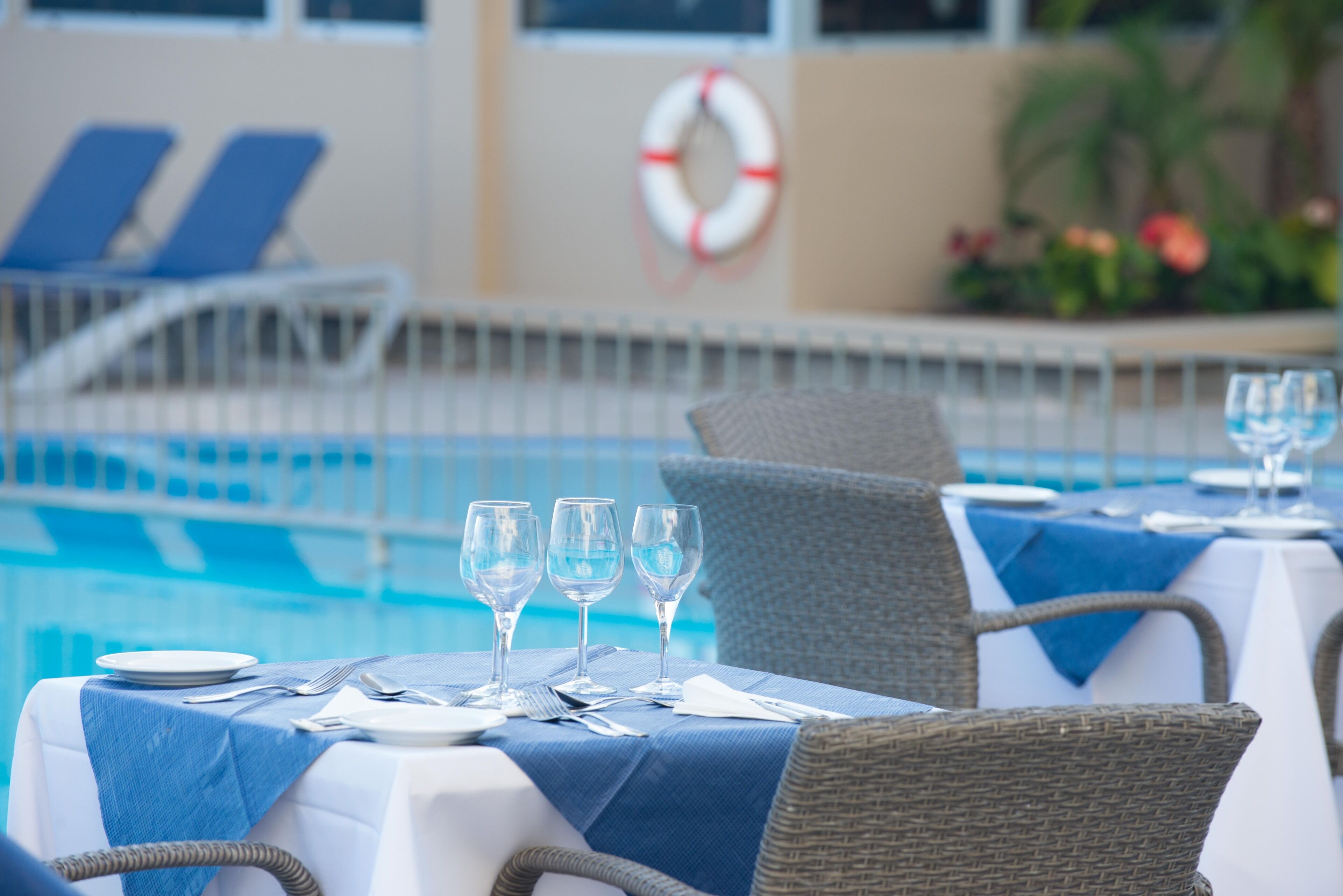 A poolside dining area with tables set for four, a lifebuoy on the wall, and lounge chairs in the background.