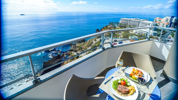A balcony with a table set for two, overlooking a coastal view with buildings and a ship in the distance.