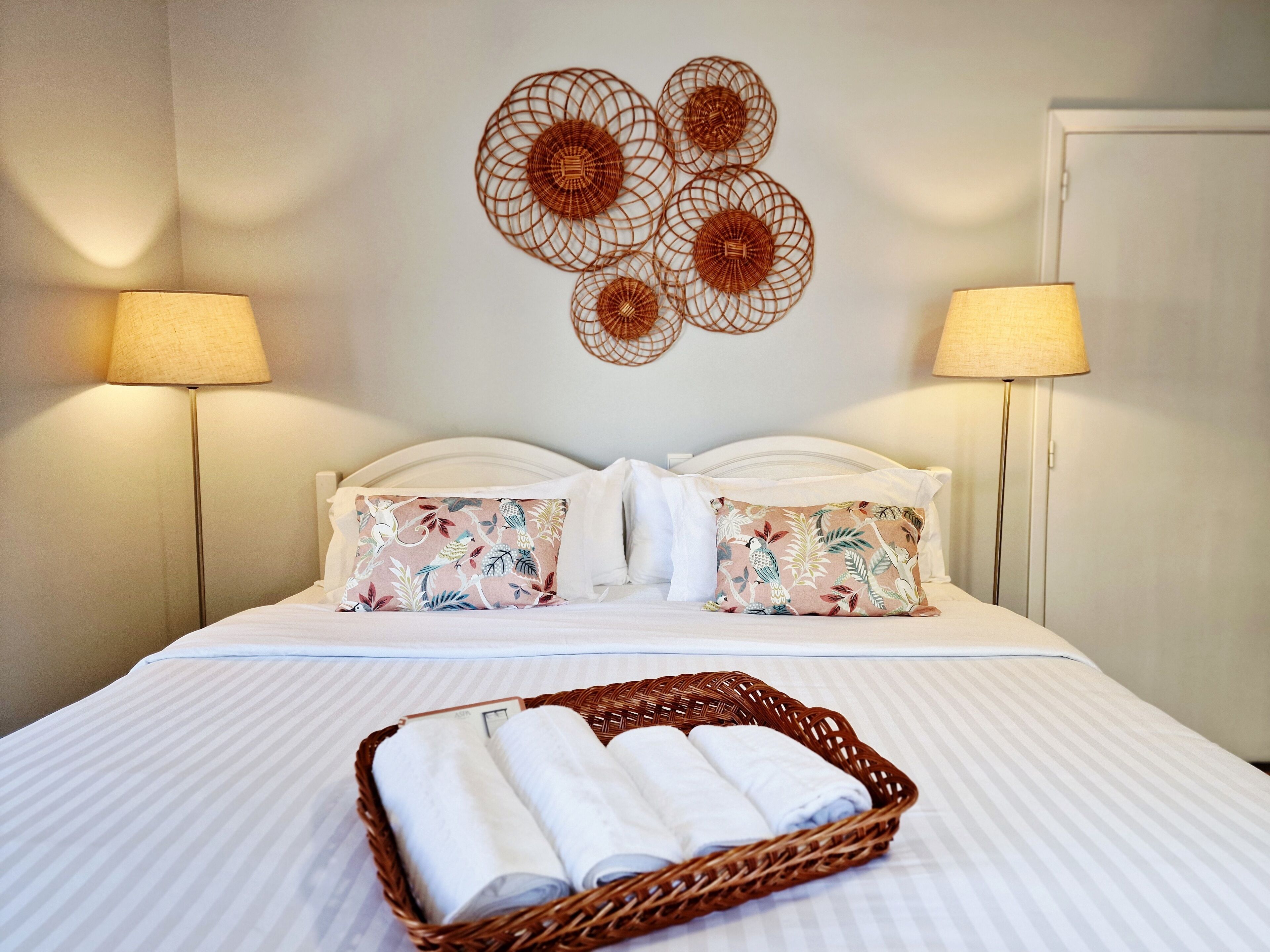 Una cama bien hecha con almohadones estampados con motivos florales, un adorno de pared tejido y una cesta con toallas dobladas.