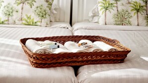 A hotel room with two beds, each with a headboard and a pillow featuring a tropical design. A wicker basket filled with rolled towels and toiletries is placed on the bed. - ASPA Studios - Urban Conscious Living (Funchal)