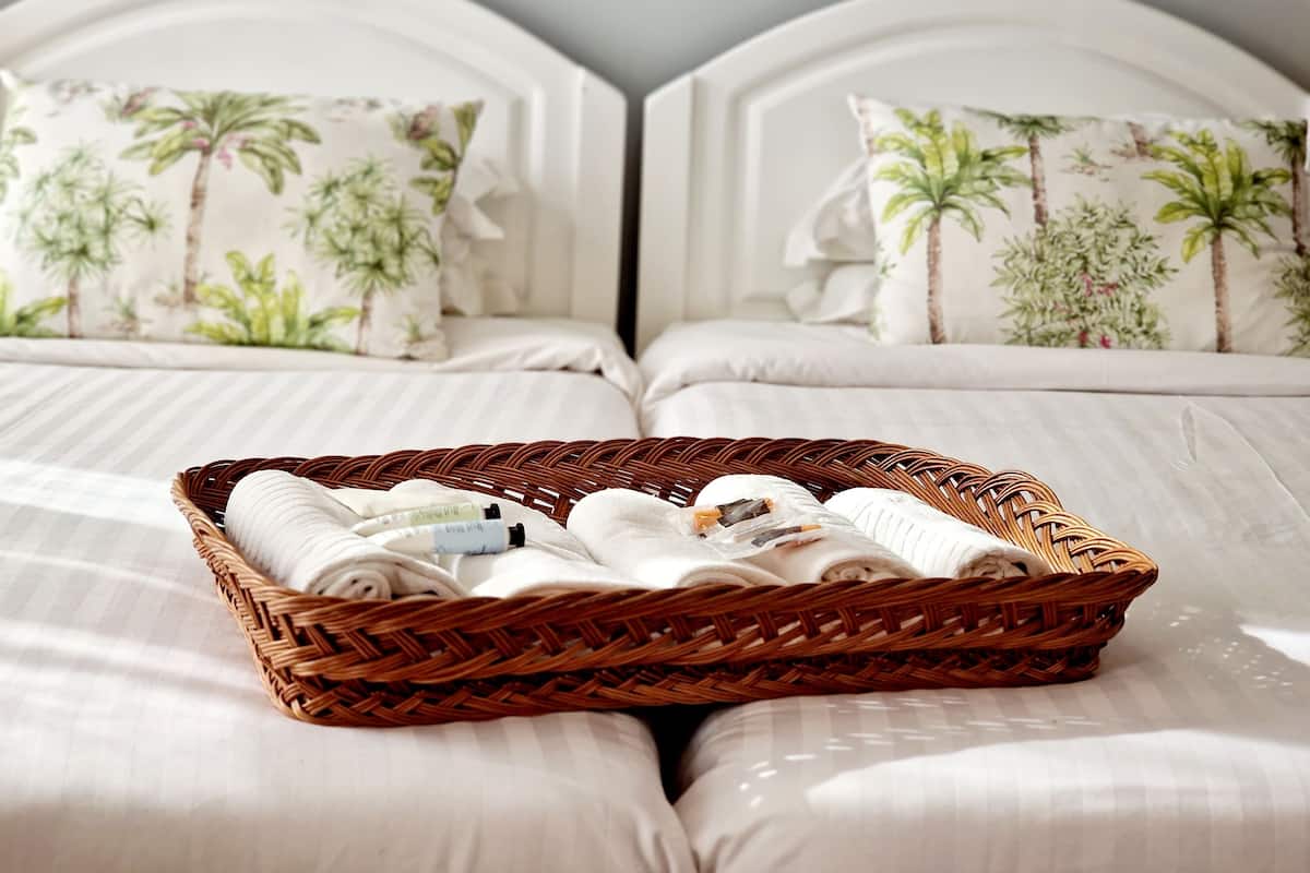 Une chambre d’hôtel avec deux lits, chacun doté d’une tête de lit et d’un oreiller orné d’un motif tropical. Un panier en osier rempli de serviettes roulées et d’articles de toilette est posé sur le lit.