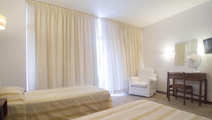 A hotel room with two beds, a desk, a chair, a TV, and a large window with curtains.