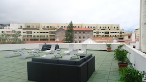 A rooftop terrace with sun loungers, a black sofa, and potted plants.