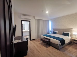 A hotel room with a bed, bedside table, and a desk with a chair. There is a window with curtains, a door, and an air conditioning unit on the wall.