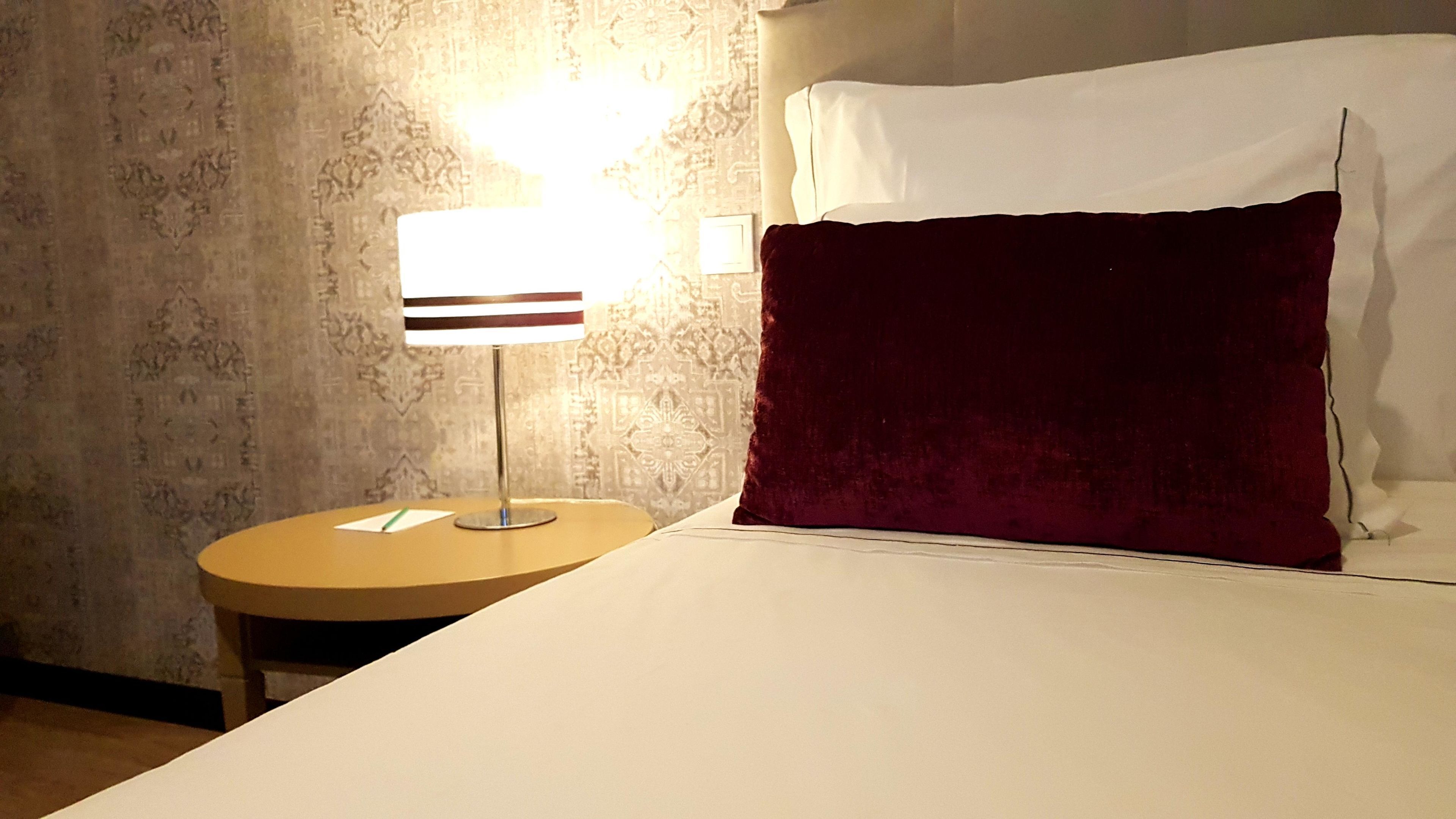 Ein Hotelzimmer mit einem Bett, einem runden Nachttisch mit Lampe, einem Wandbild mit Muster und einem roten Zierkissen.