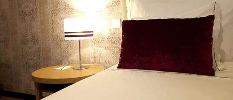 Ein Hotelzimmer mit einem Bett, einem runden Nachttisch mit Lampe, einem Wandbild mit Muster und einem roten Zierkissen.