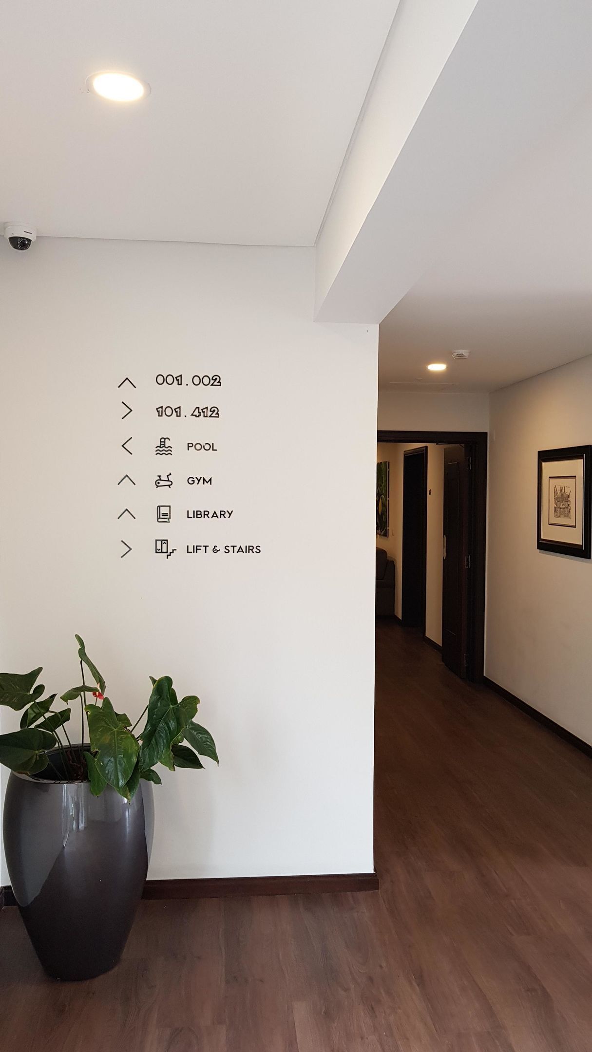 A hallway with a white wall featuring directional signs to various amenities like the pool, gym, library, and lift & stairs.