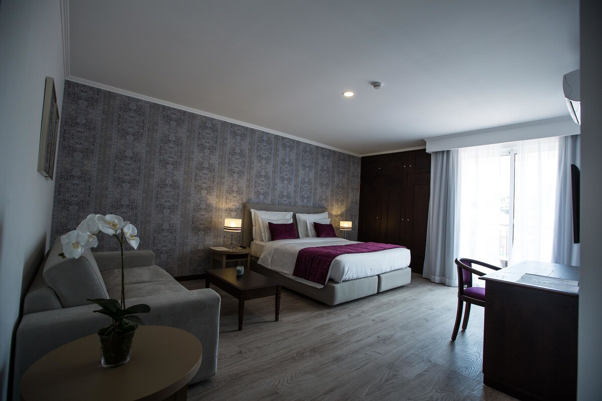 A modern hotel room with a bed, a desk, a chair, a small table, and a potted plant.
