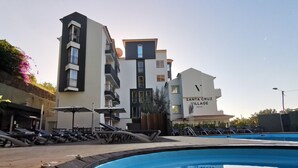 Een zwembadgedeelte met ligbedden en parasols, een modern gebouw met de tekst 'Santa Cruz Village Hotel', en een heldere lucht.
