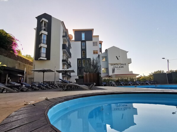 A pool area with lounge chairs and umbrellas, a modern building labeled "Santa Cruz Village Hotel," and a clear sky.
