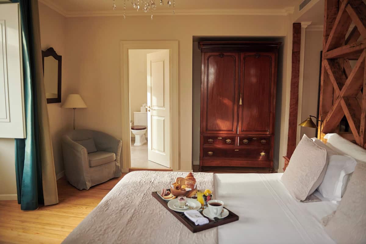 A hotel room with a large bed, a wooden wardrobe, a sitting area with a chair, and a breakfast tray on the bed.