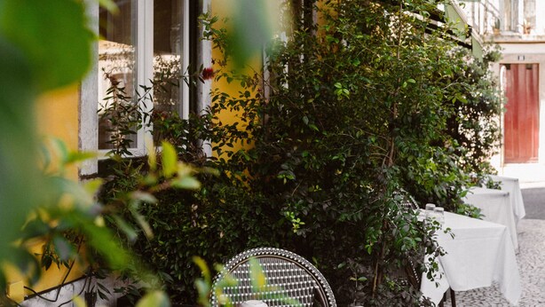 An outdoor dining area with tables set for two, surrounded by greenery and a building facade.
