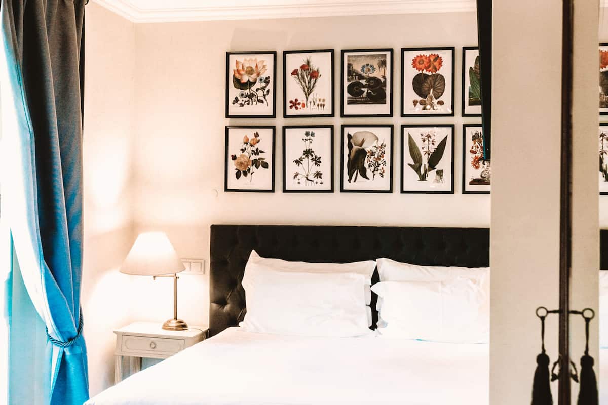 A neatly made bed with white linens, a dark headboard, and a series of framed floral artworks on the wall above.