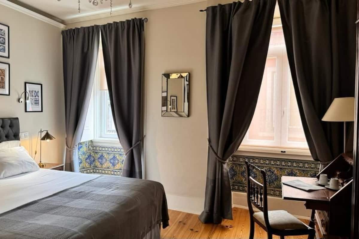 A bedroom with a bed, a desk, and a chair. There are curtains, a mirror, and framed pictures on the wall.