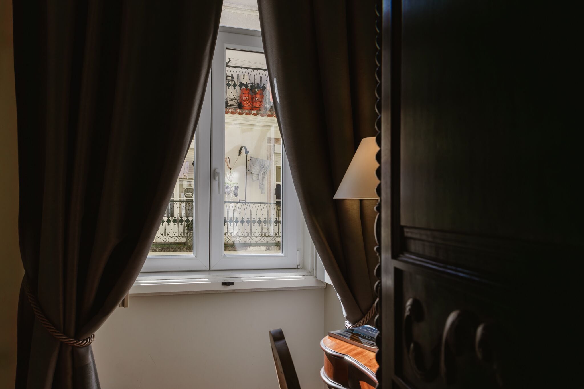 A room with a window, curtains, a lamp, and a view of a balcony with a railing and potted plants.