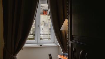 A room with a window, curtains, a lamp, and a view of a balcony with a railing and potted plants.