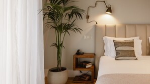 A bedroom with a bed, a bedside table with books, a lamp, and a potted plant.