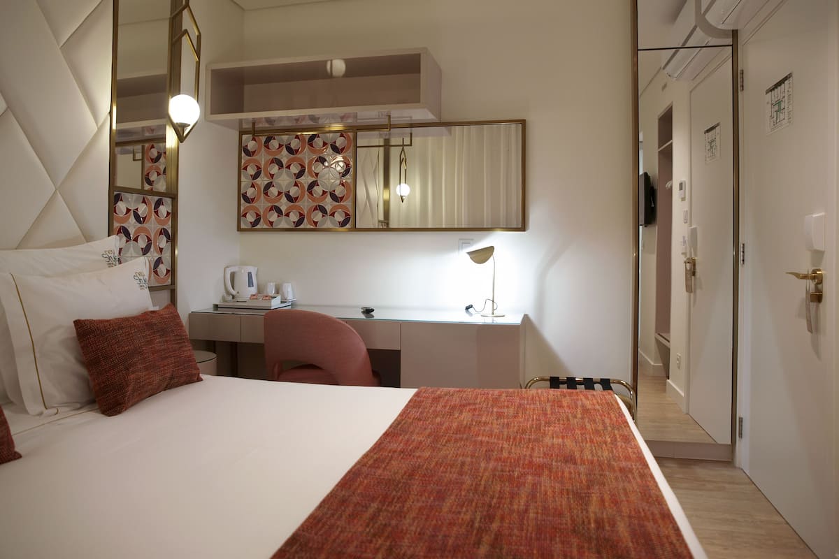 Quarto de hotel com uma cama grande, uma escrivaninha, uma cadeira, um espelho e um guarda-roupa.