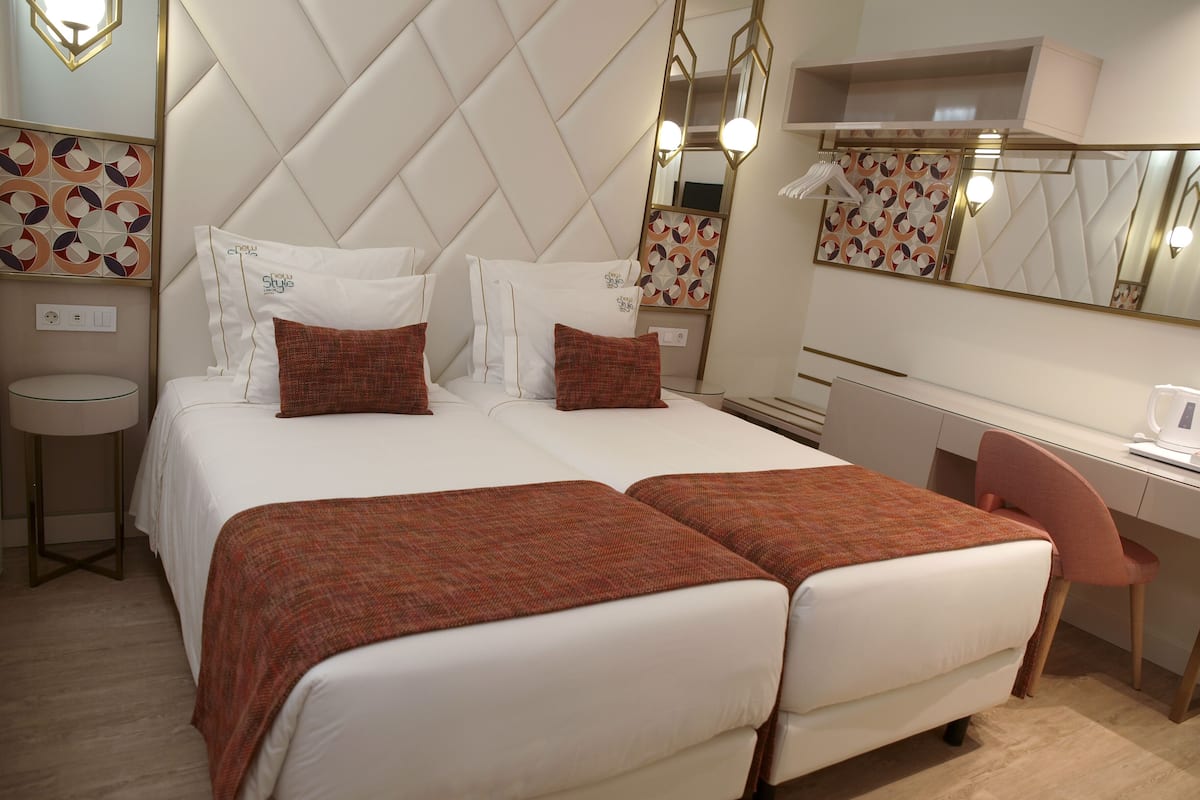Quarto de hotel com uma cama grande, uma escrivaninha e uma cadeira. Há um espelho sobre a cama e uma pequena mesa com abajur.