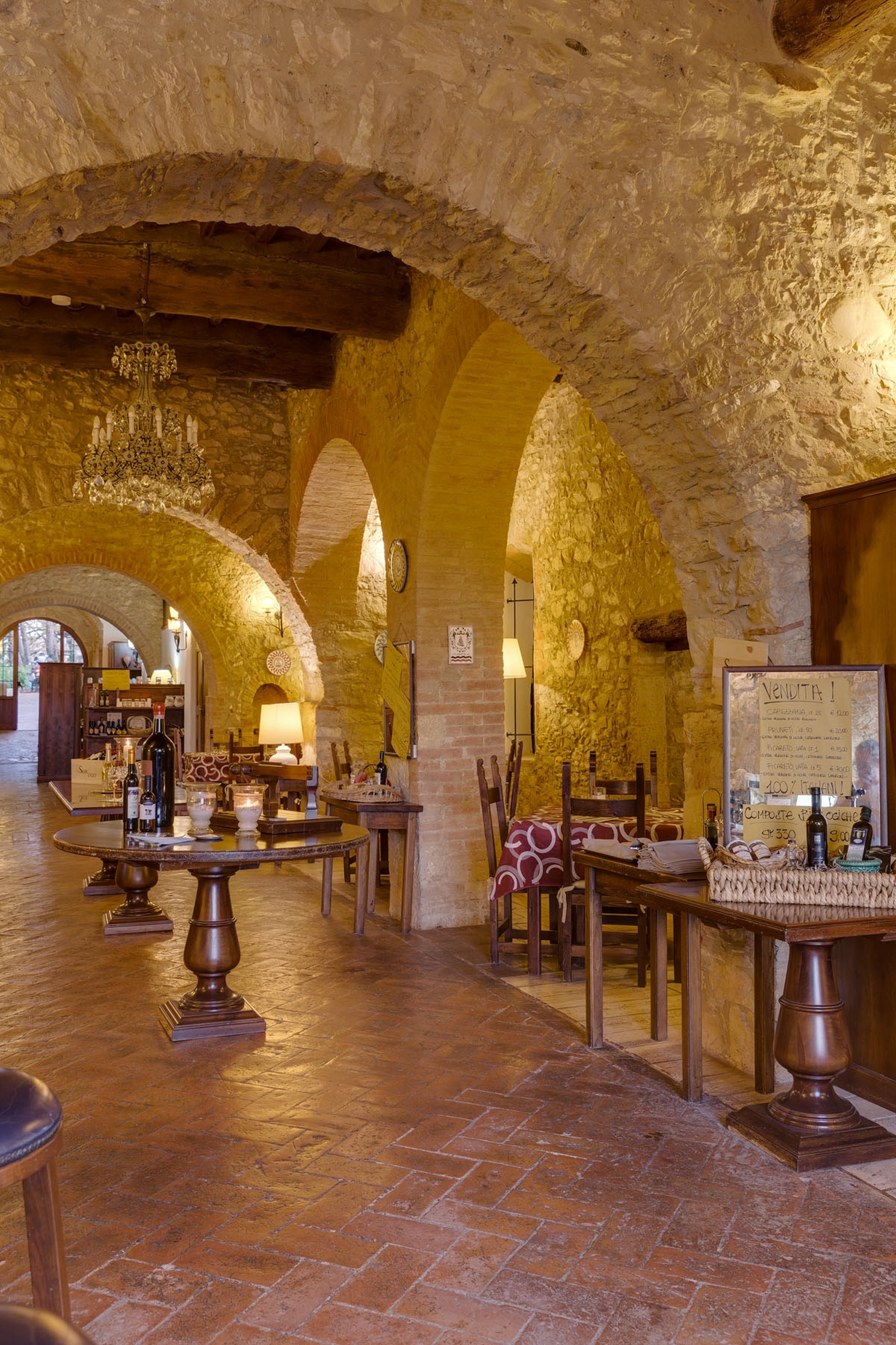 Un interno rustico con archi in pietra, arredi in legno e un lampadario. I tavoli sono apparecchiati con bottiglie di vino e bicchieri.