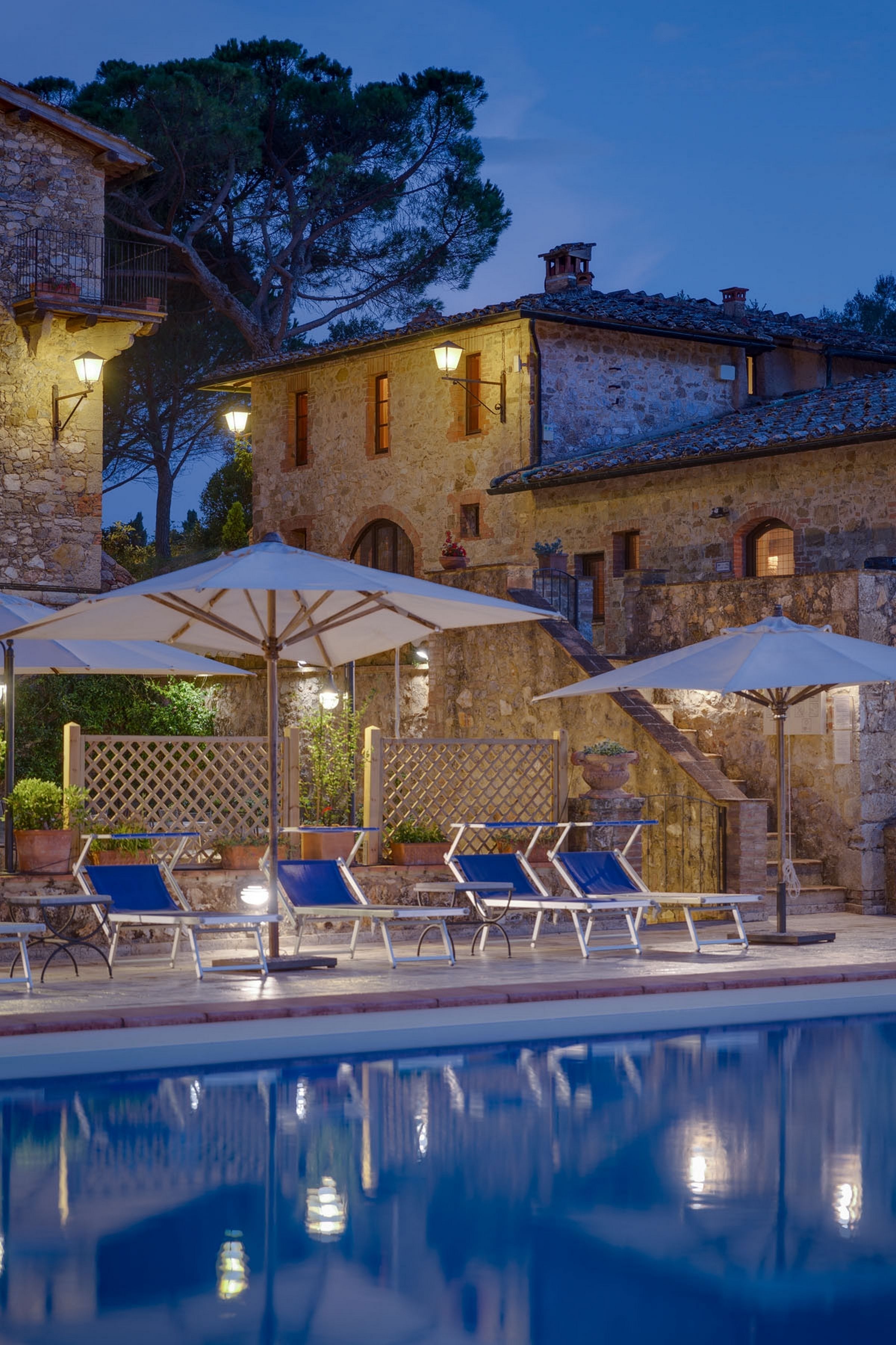A pool area with lounge chairs and umbrellas, stone buildings in the background, and a clear pool reflecting the surroundings.