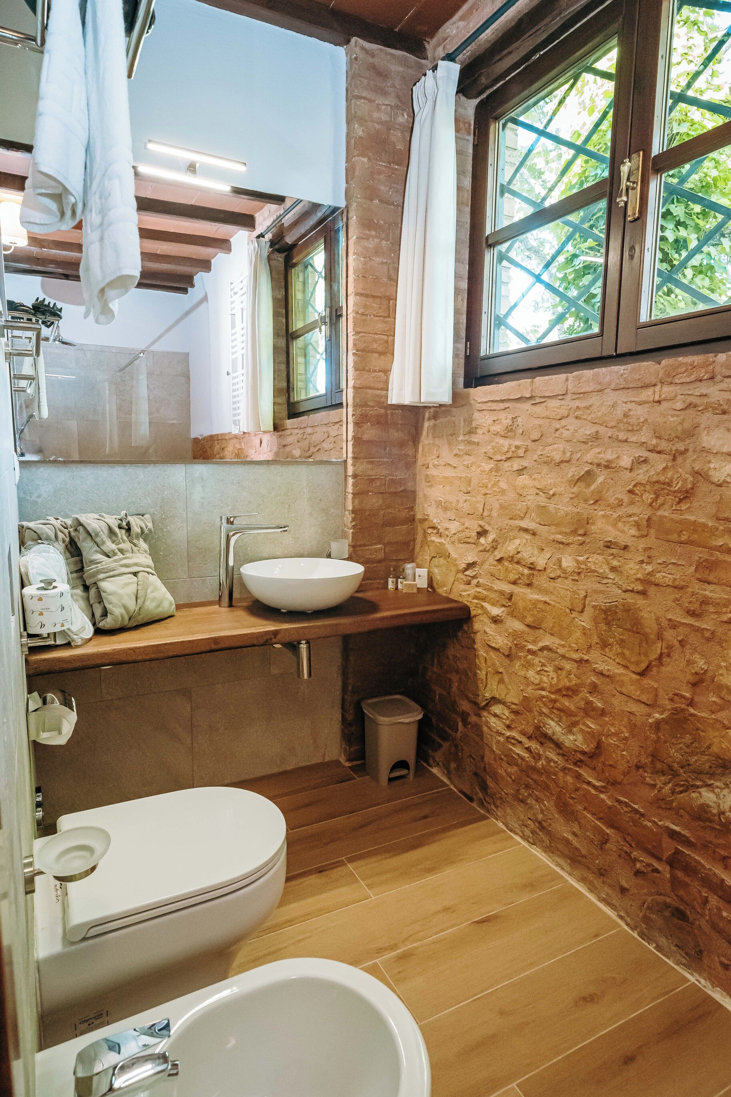 A bathroom with a stone wall, wooden floor, a white sink, a toilet, and a window with a view of greenery.