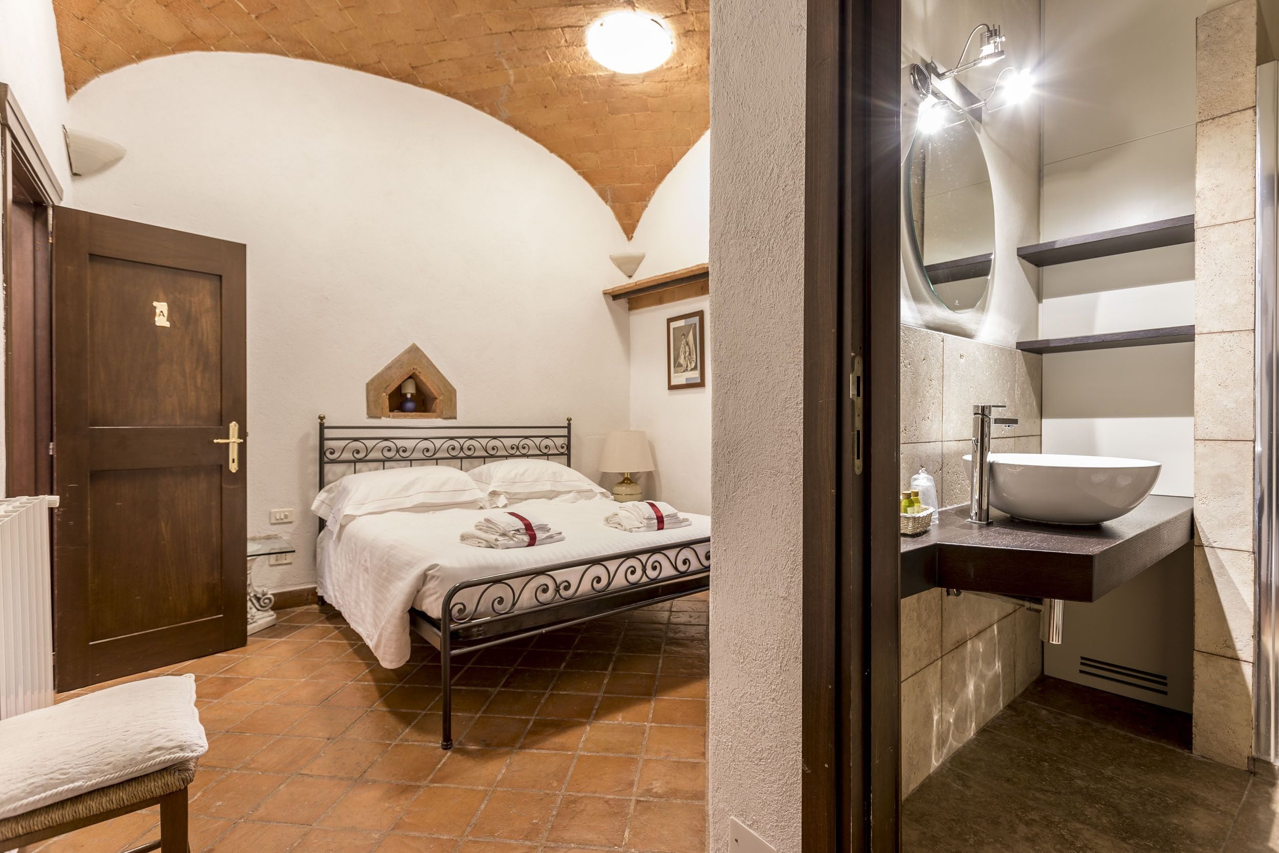 Una camera da letto con un letto, una porta in legno, una panca e un bagno con lavandino e specchio.