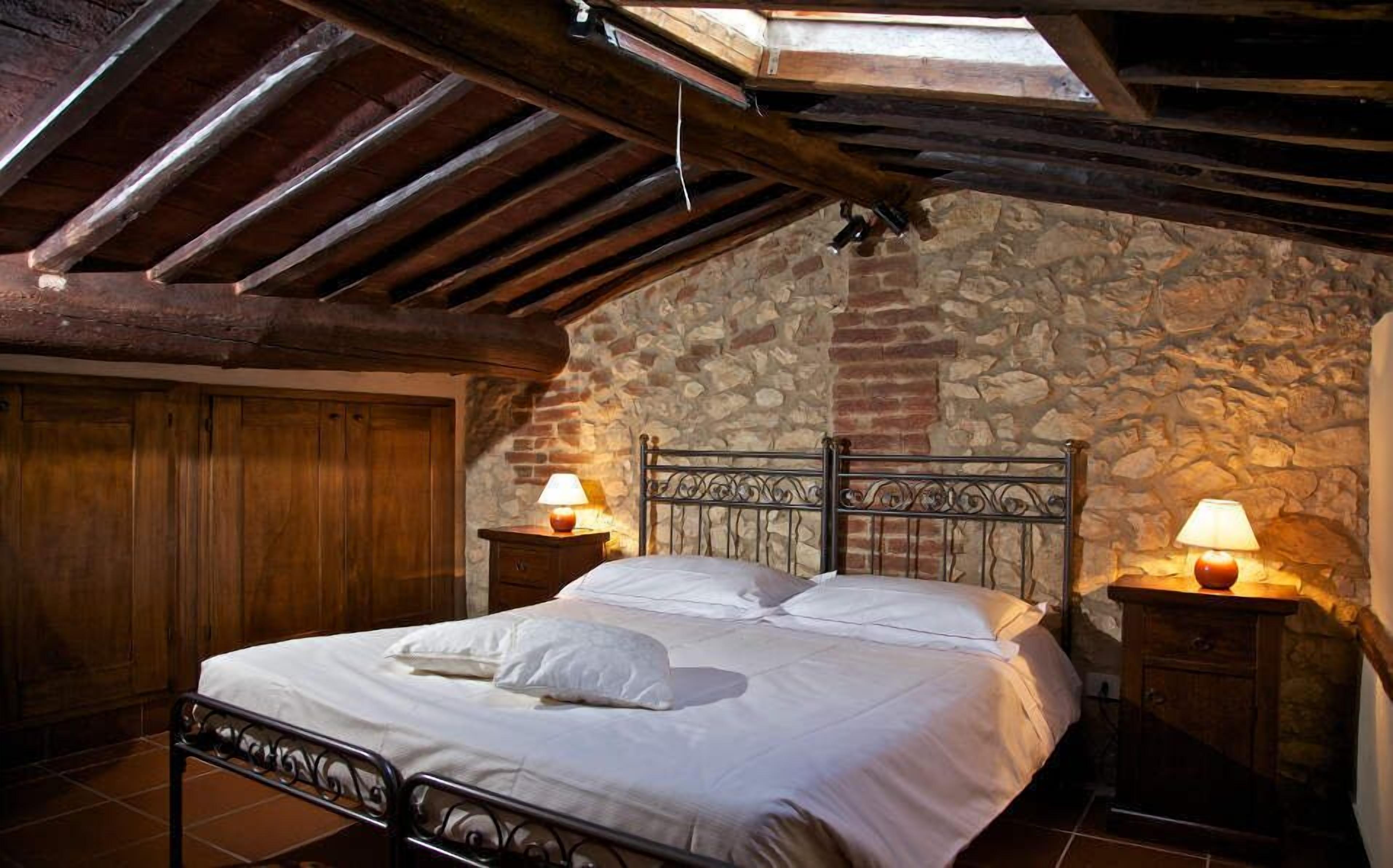 A bedroom with a stone wall, wooden ceiling beams, a metal bed frame, and two bedside lamps.