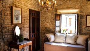 Ein gemütlicher Raum mit Steinwand, Holztür, einem Korbsessel mit Kissen, einem kleinen Tisch mit Spiegel und Vase sowie einer Kristallleuchter.