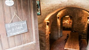 Ein rustikaler Innenraum mit einem Steingewölbe, das zu einem Essbereich führt. Ein Kreidetafel-Schild an der Wand zeigt: "COLAZIONE 8-10 19:00 Buon Appetito!"