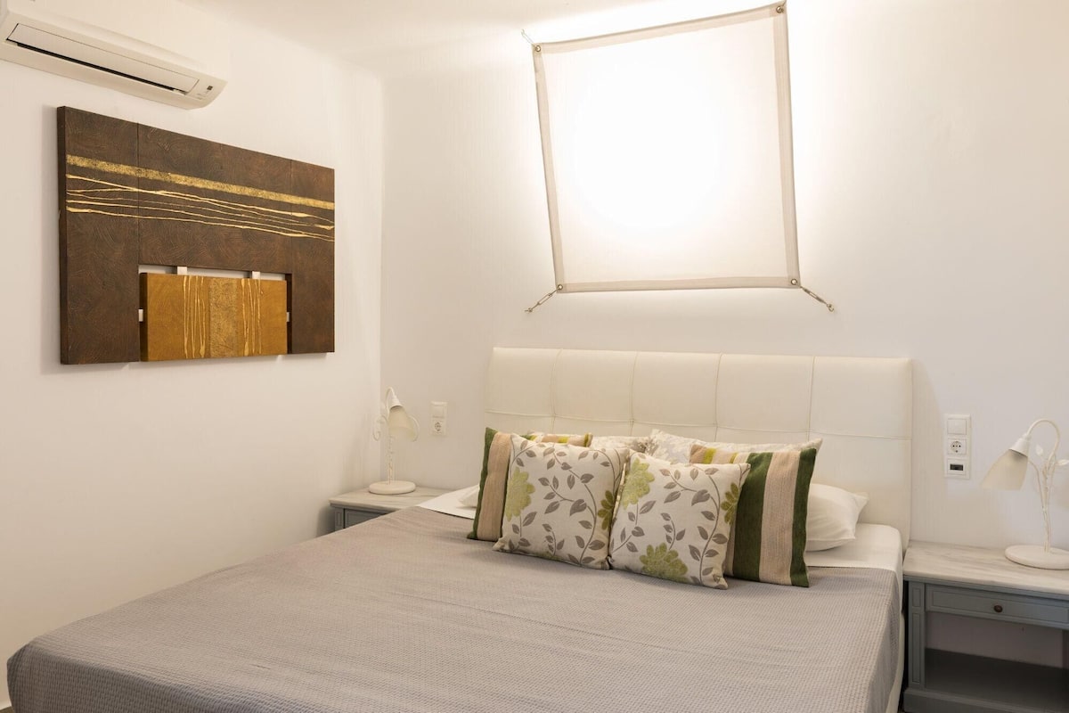 Un dormitorio con cama, mesitas de noche, aire acondicionado de pared y ventana con marco blanco.