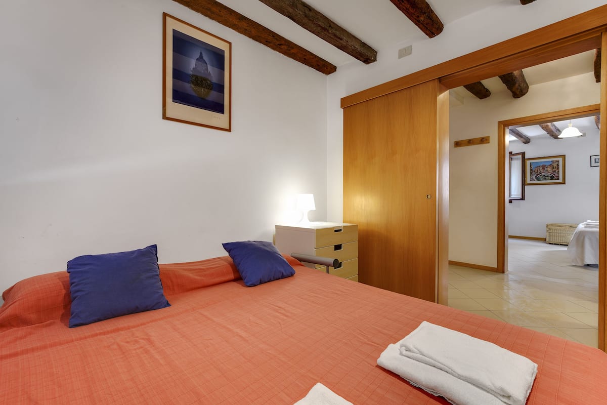 Un dormitorio con cama, almohadas, una mesita de noche, una lámpara y un armario de madera.