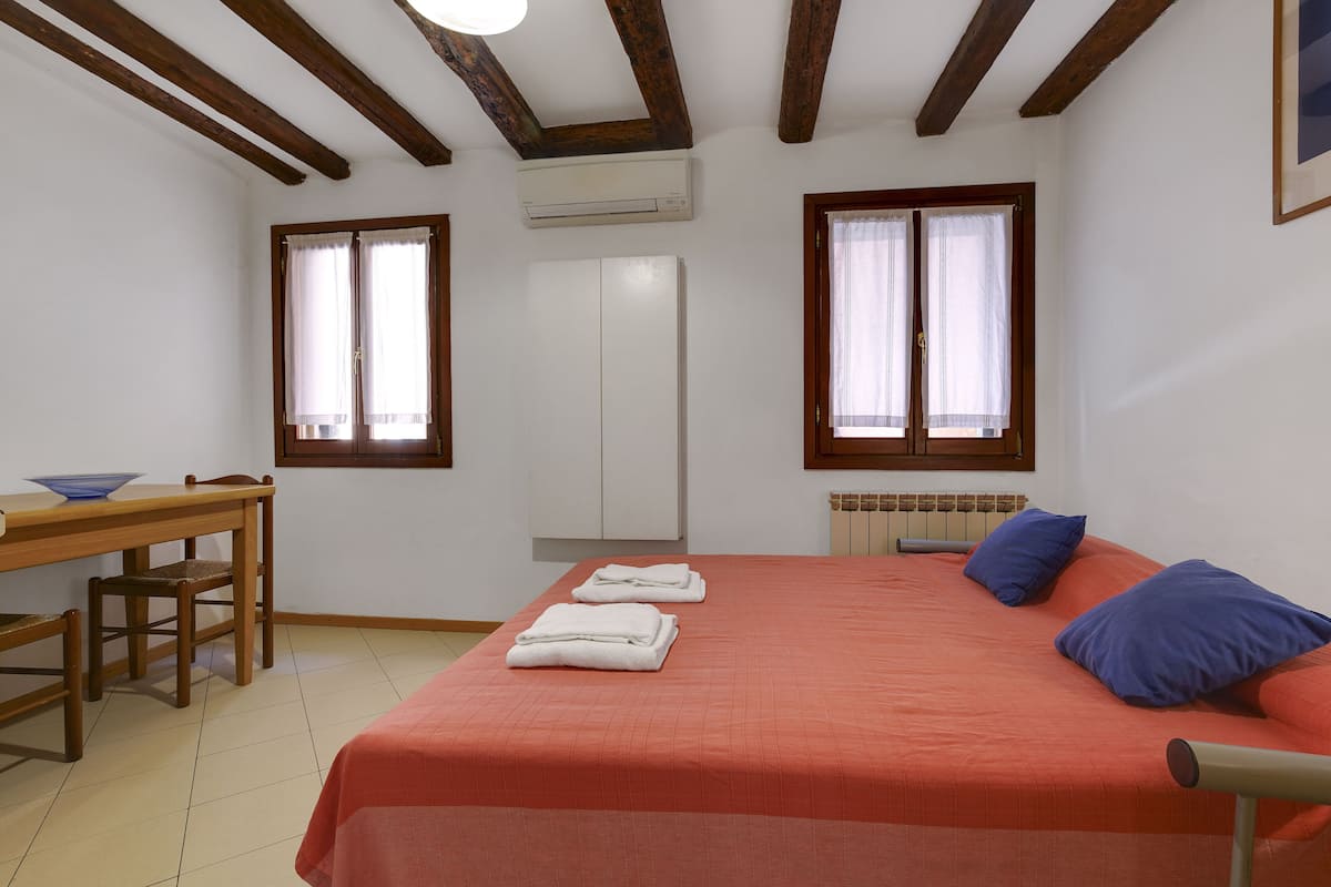 Un dormitorio con una cama, dos ventanas, un radiador, una mesa de madera con una silla y un armario blanco.
