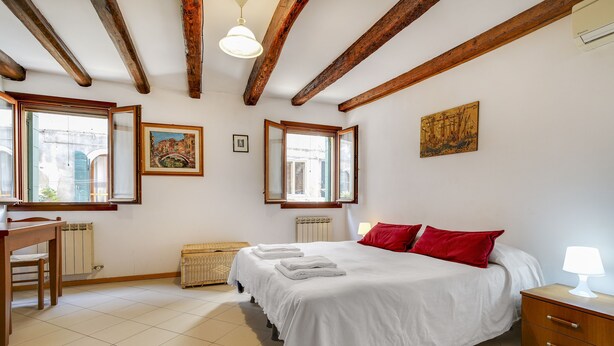 A bedroom with a large bed, wooden beams, a desk, and a window with a view of a building. - Bed & Breakfast Giardini (Venice)