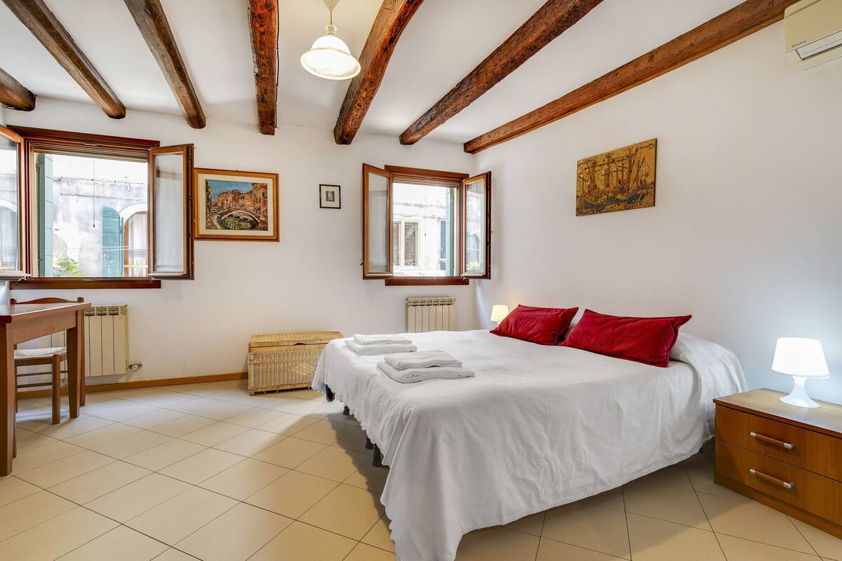 Un dormitorio con una cama grande, vigas de madera, un escritorio y una ventana con vista a un edificio.
