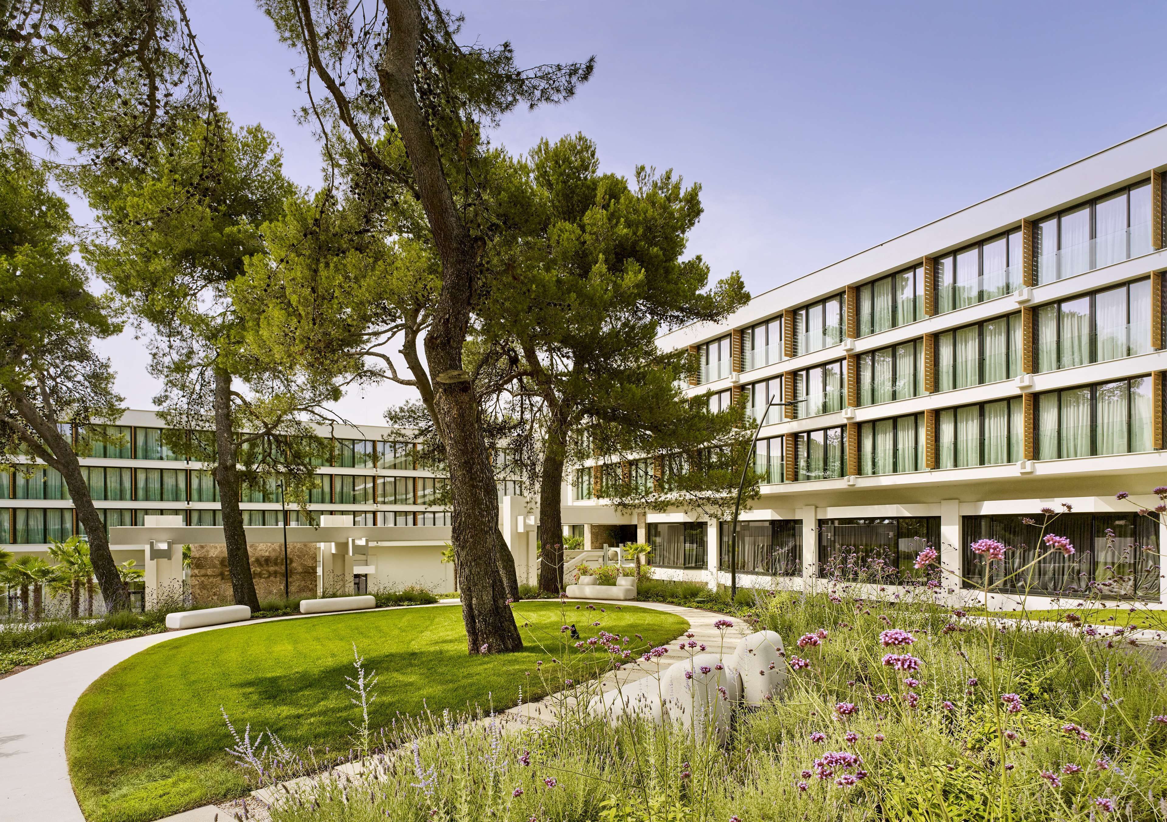 A modern building with multiple windows and a well-maintained garden with trees and flowers.