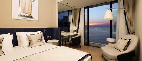 Habitación de hotel con una cama grande, zona de estar con una mesa y sillas, vista al océano y un cuadro en la pared.