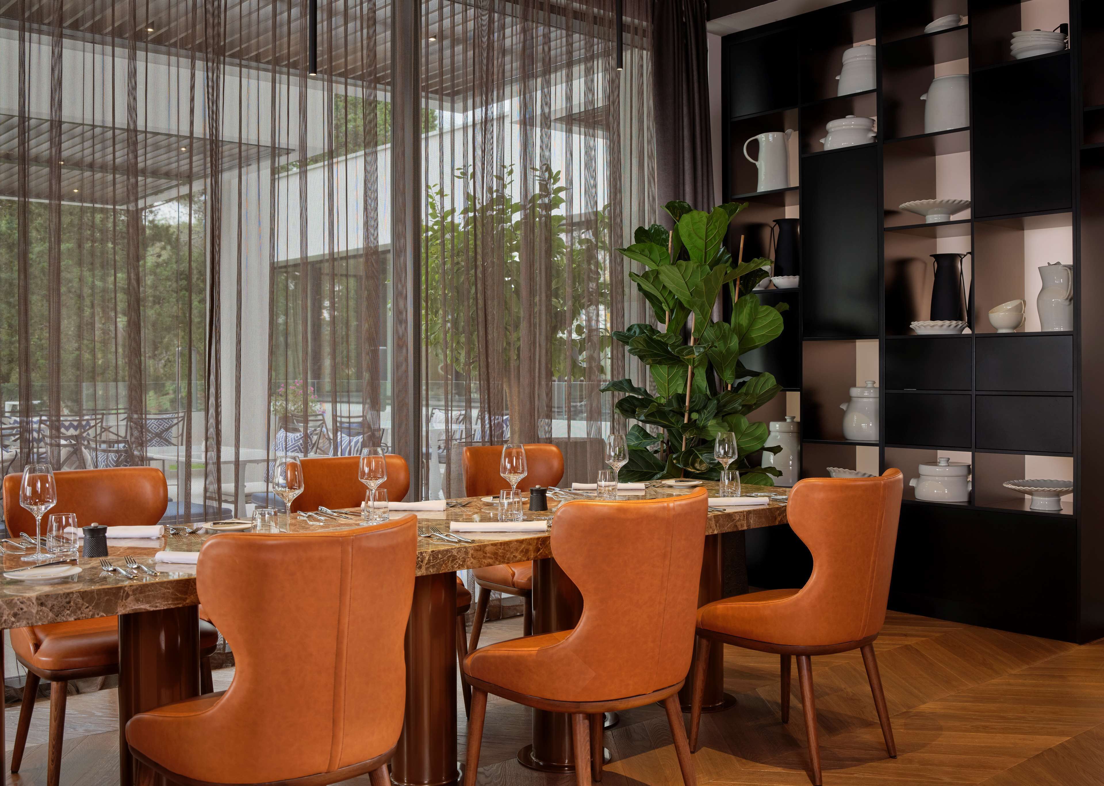 Un comedor con una mesa preparada para cuatro personas, sillas naranjas alrededor y una planta grande en maceta al fondo.