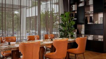 Un coin repas avec une table dressée pour quatre personnes, entourée de chaises orange, et une grande plante en pot en arrière-plan.