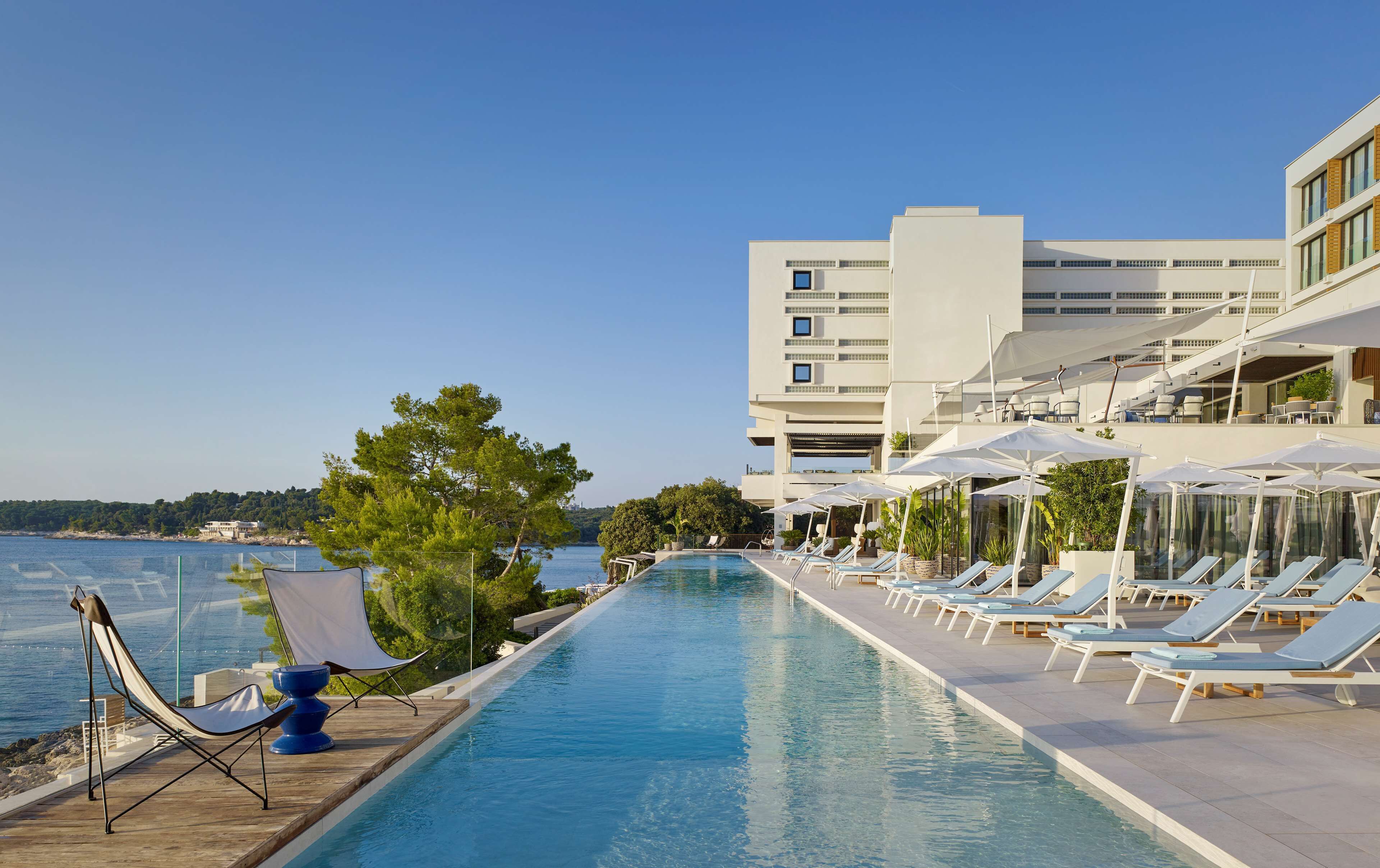 Un espace aménagé au bord de la piscine, avec des chaises longues et des parasols, une vue dégagée sur la mer et un bâtiment moderne à plusieurs étages.