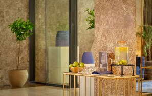A reception area with a marble wall, a glass door, a reception desk with a bowl of green apples, a glass dispenser with a yellow drink, and a welcome sign.