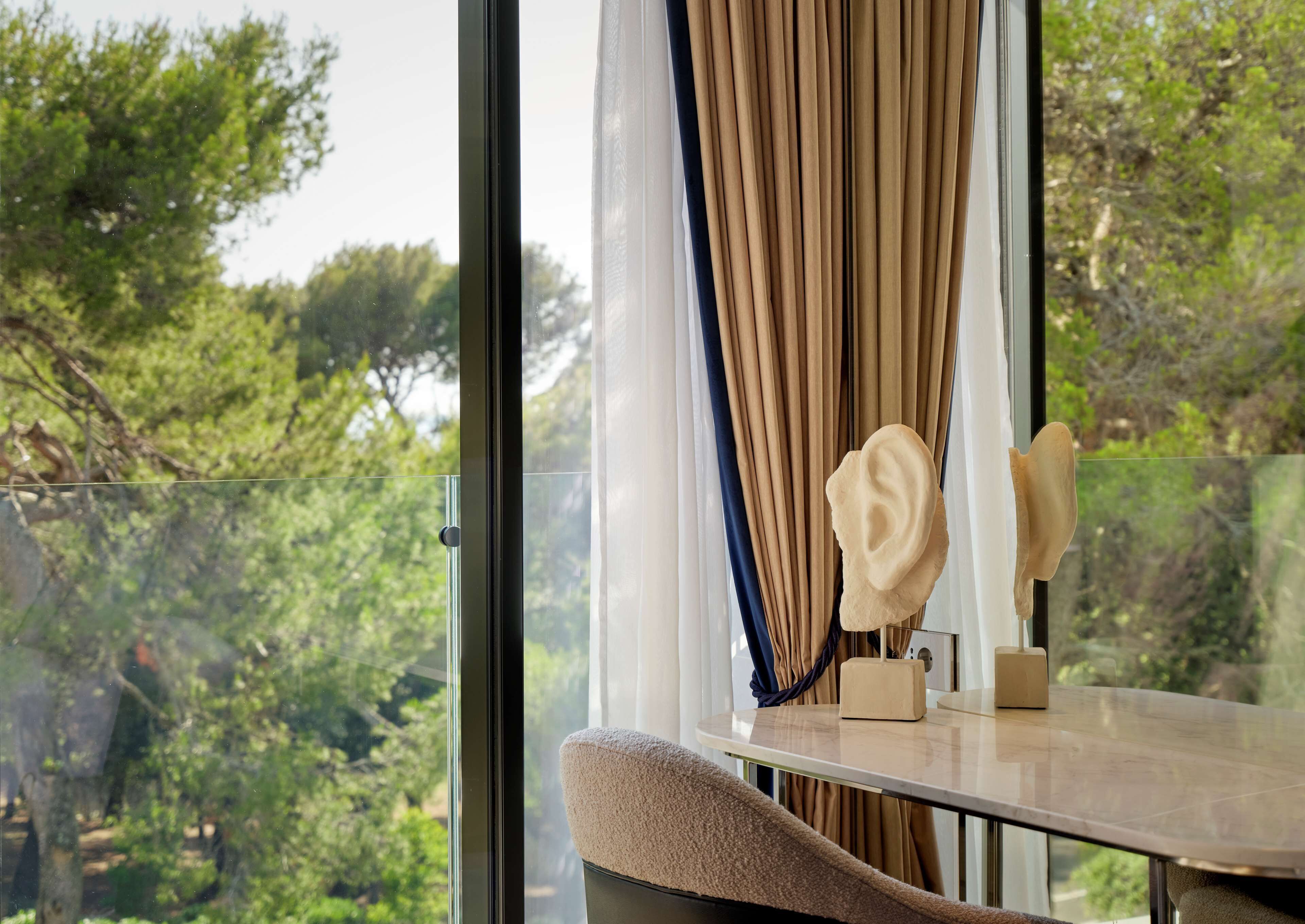 A room with a large window, a view of greenery outside, a table with a vase and a small box, and a cushioned chair.