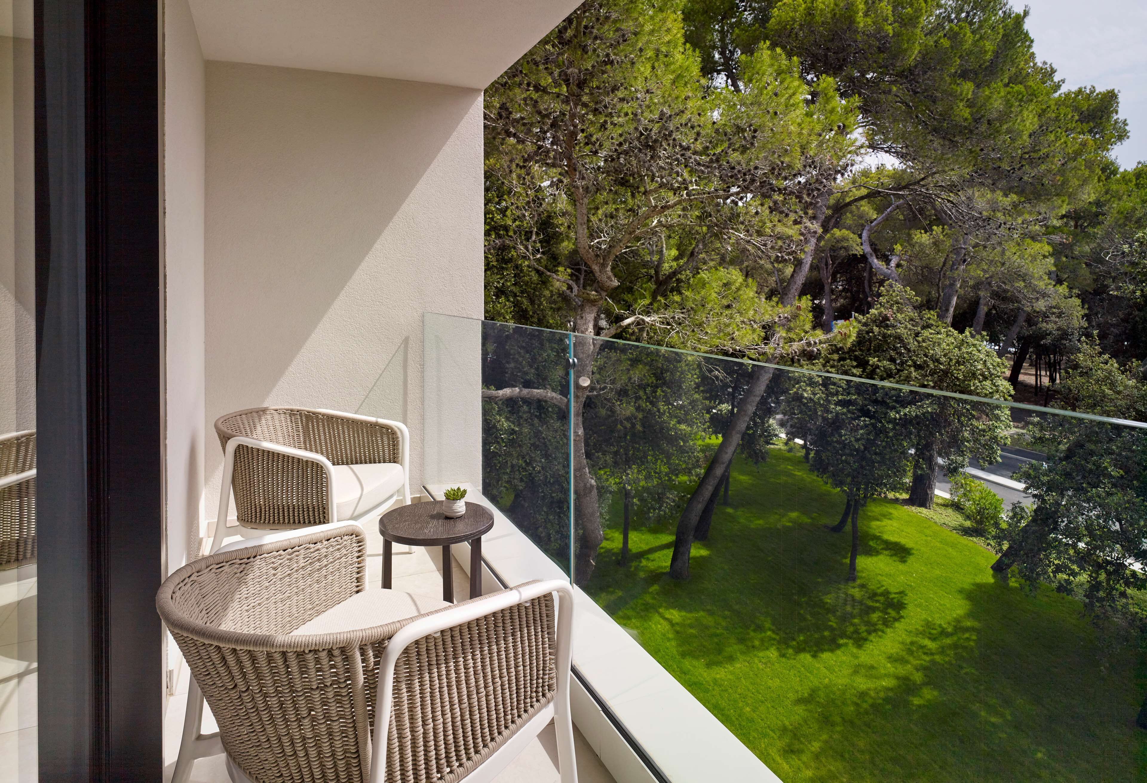 A balcony with wicker furniture and a small table, overlooking a well-maintained garden with trees and grass.