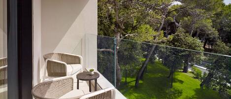 A balcony with wicker furniture and a small table, overlooking a well-maintained garden with trees and grass.